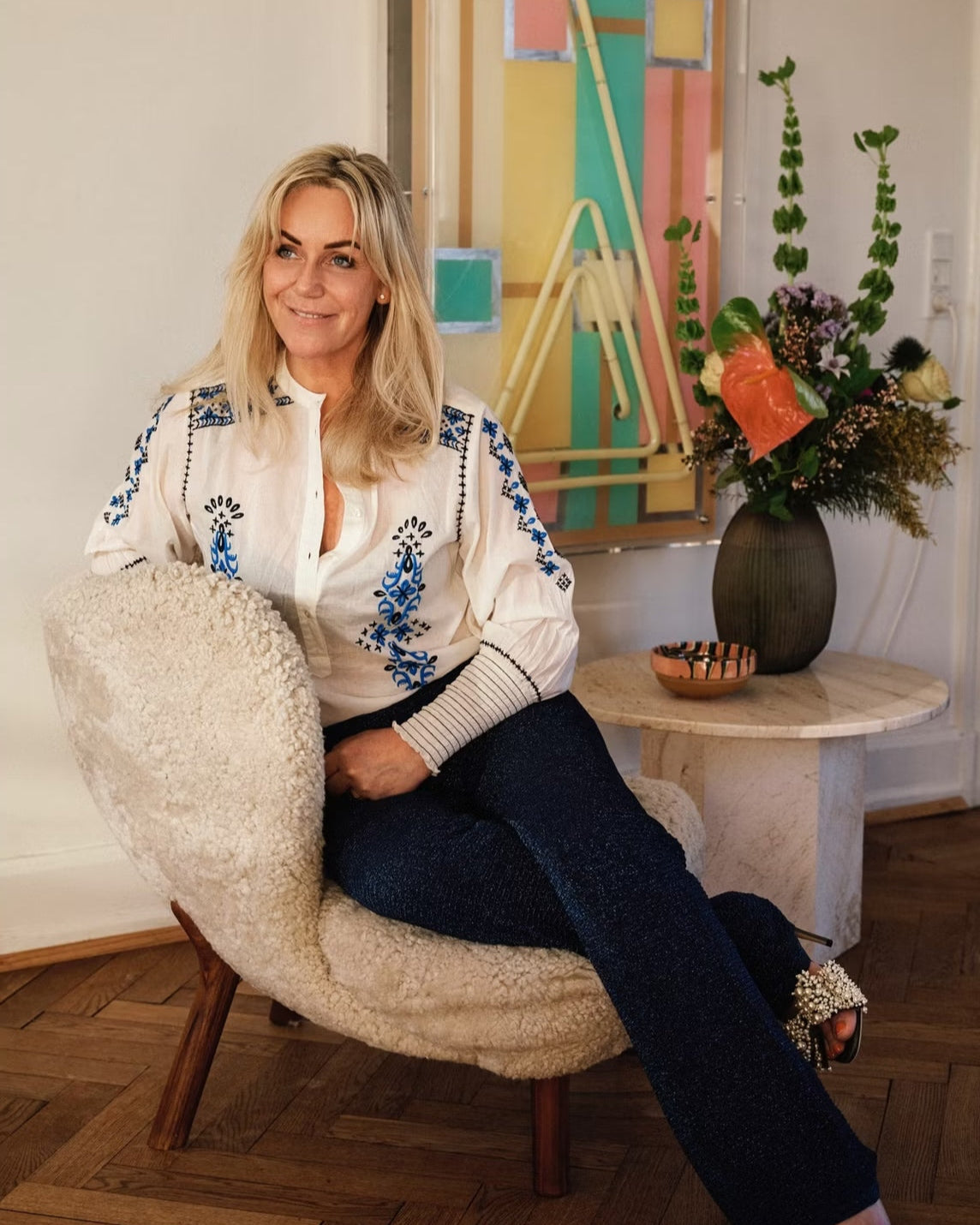 Woman sitting on a textured chair in a room with a colorful abstract painting and floral arrangement.