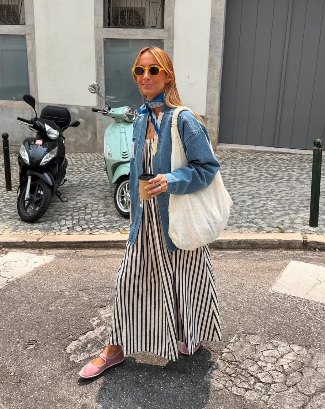 Woman in a striped dress and denim jacket standing on a street with a scooter in the background.